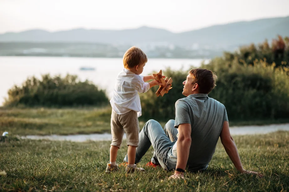A father sitting on the grass by a lake, watching his young son hold up a wooden toy plane.